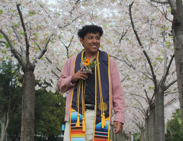 Alexander in graduation stole and honor cords, holding flowers in front of cherry blossoms.