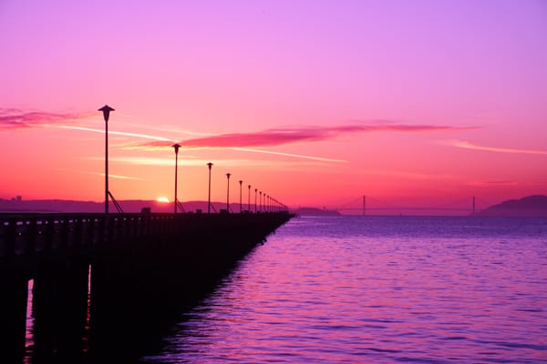 A pier extending into the horizon, where it meets the Golden Gate bridge, with the sun setting in the background